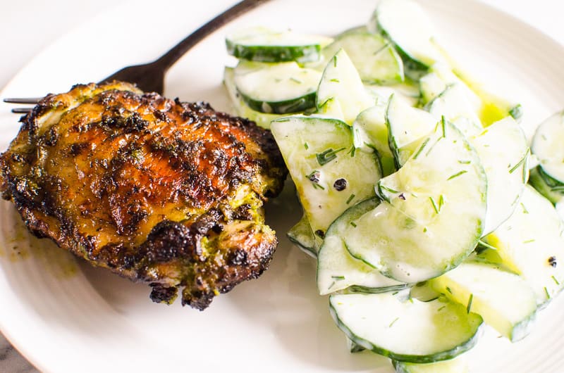 Grilled chicken thighs served with cucumber salad on white plate and black fork.