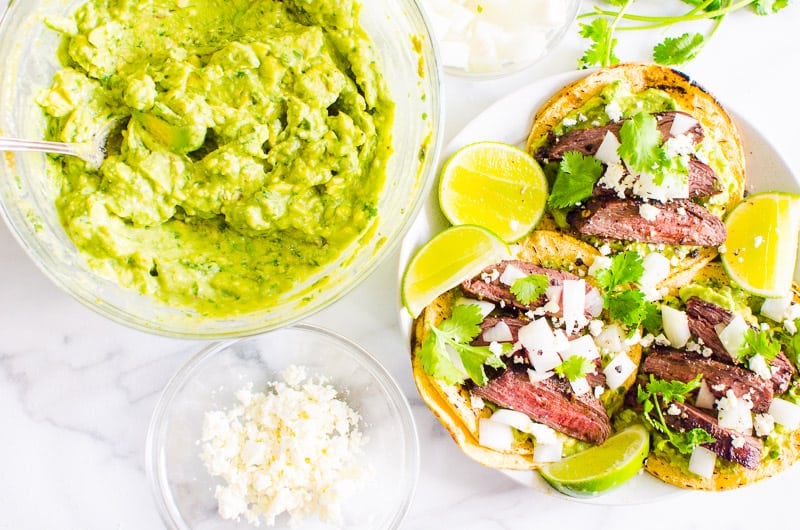 Guacamole in a bowl, steak tacos on plate and crumbled cheese in a bowl.