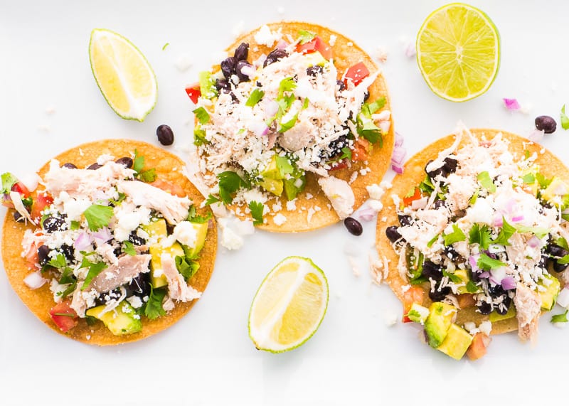 Three chicken tostadas with avocado, beans, cheese and lime wedges around on a countertop.