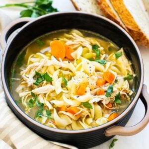 Turkey noodle soup in a bowl with parsley garnish. Sliced bread and linen on counter.