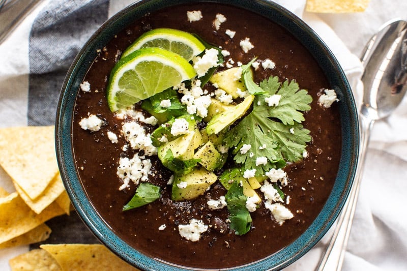 Black bean soup garnished with avocado, feta cheese, lime and cilantro in a bowl.