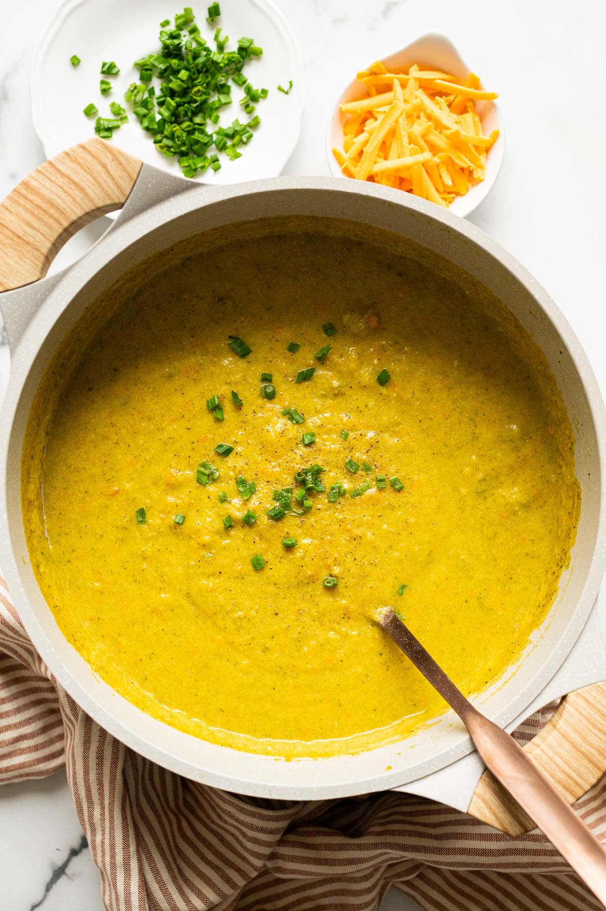 Healthy broccoli cheddar soup garnished with chives in a pot with ladle. Cheese, chives and linen towel on a counter.