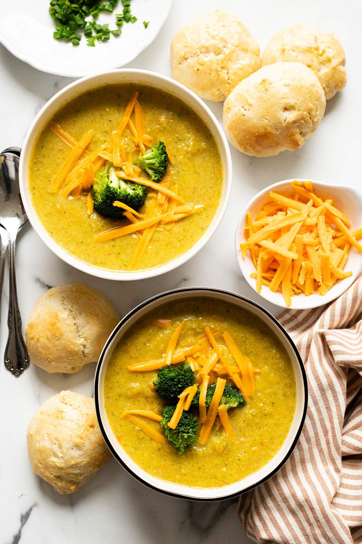 Two bowls with broccoli cheddar soup. Spoons, dinner rolls, cheese, chives and napkin on the countertop.