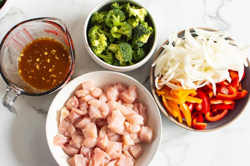 Cubed chicken, chopped onions and bell pepper, broccoli florets and stir fry sauce in separate bowls.