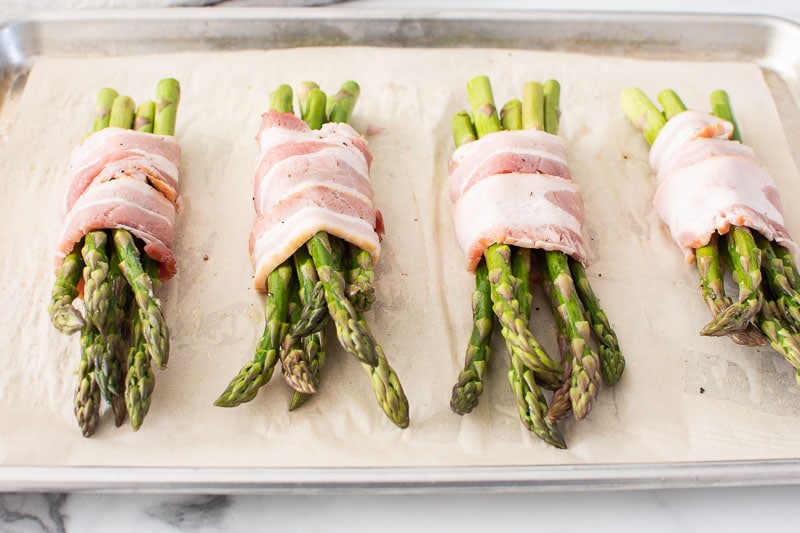 four asparagus bunches wrapped in bacon on baking sheet