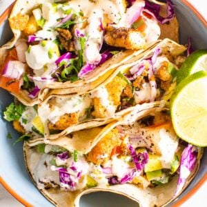 Three fish tacos with taco sauce and fresh limes in blue bowl.