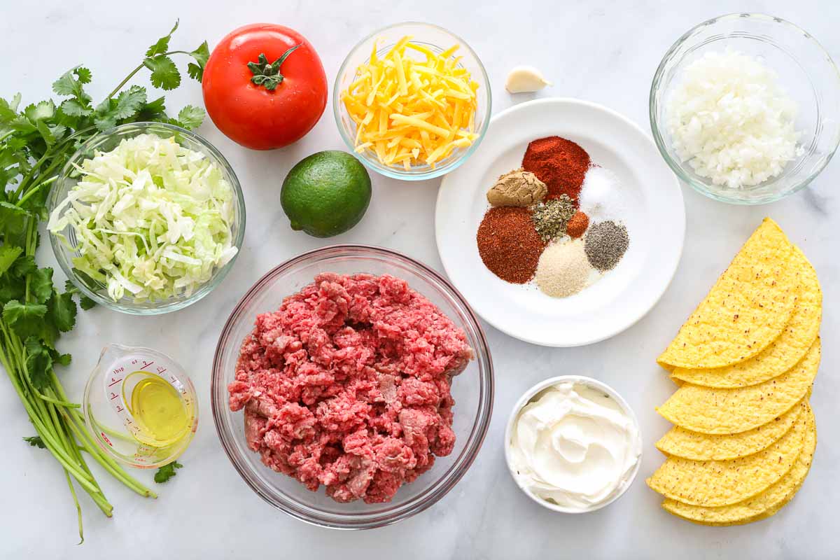 Ingredients for Ground Beef Tacos, including beef, cheese, taco shells, seasonings, sour cream, onion, garlic, tomatoes, lettuce, cilantro, lime, and oil.