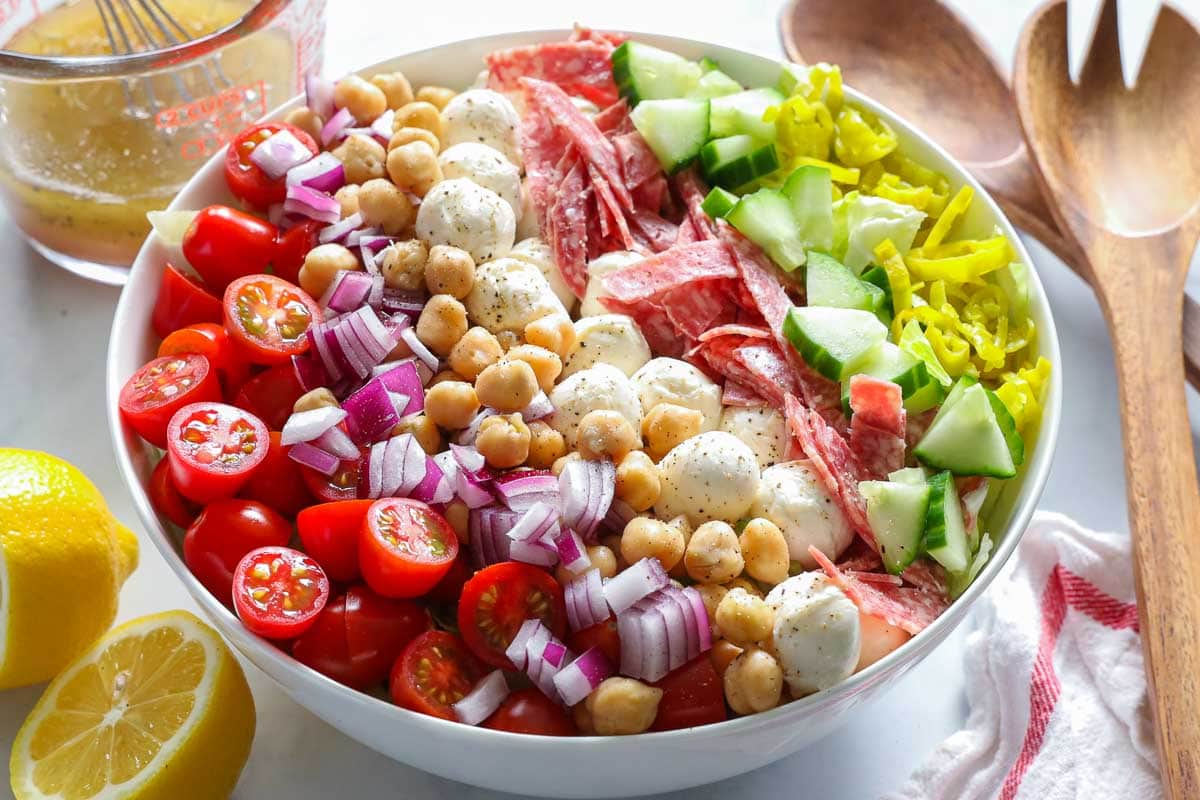 Assembled Italian chopped salad in a bowl with serving spoons and Italian dressing in measuring cup.