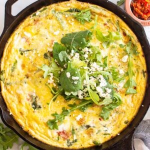 Vegetable frittata in skillet with feta and greens on top.