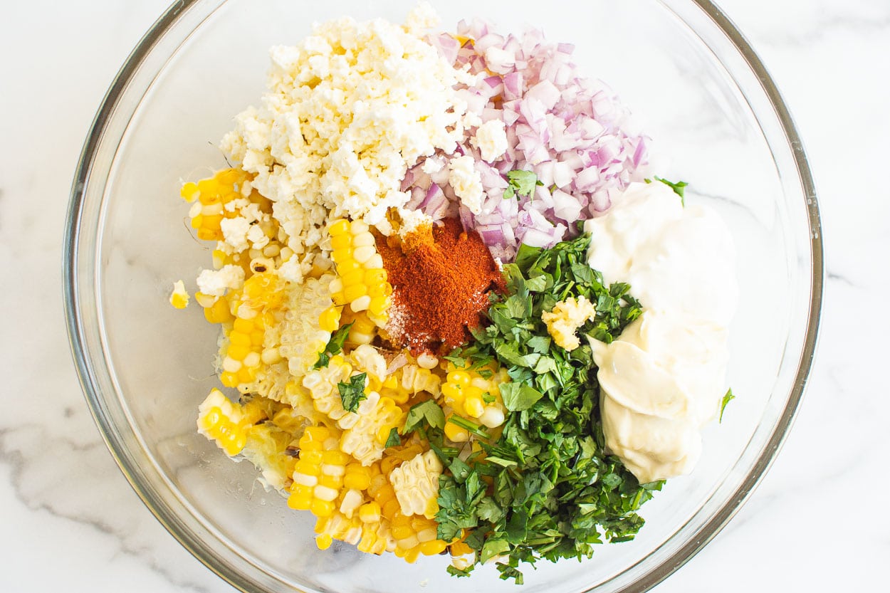 Cooked corn, cilantro, red onion, feta, mayo, yogurt and spices in glass bowl.
