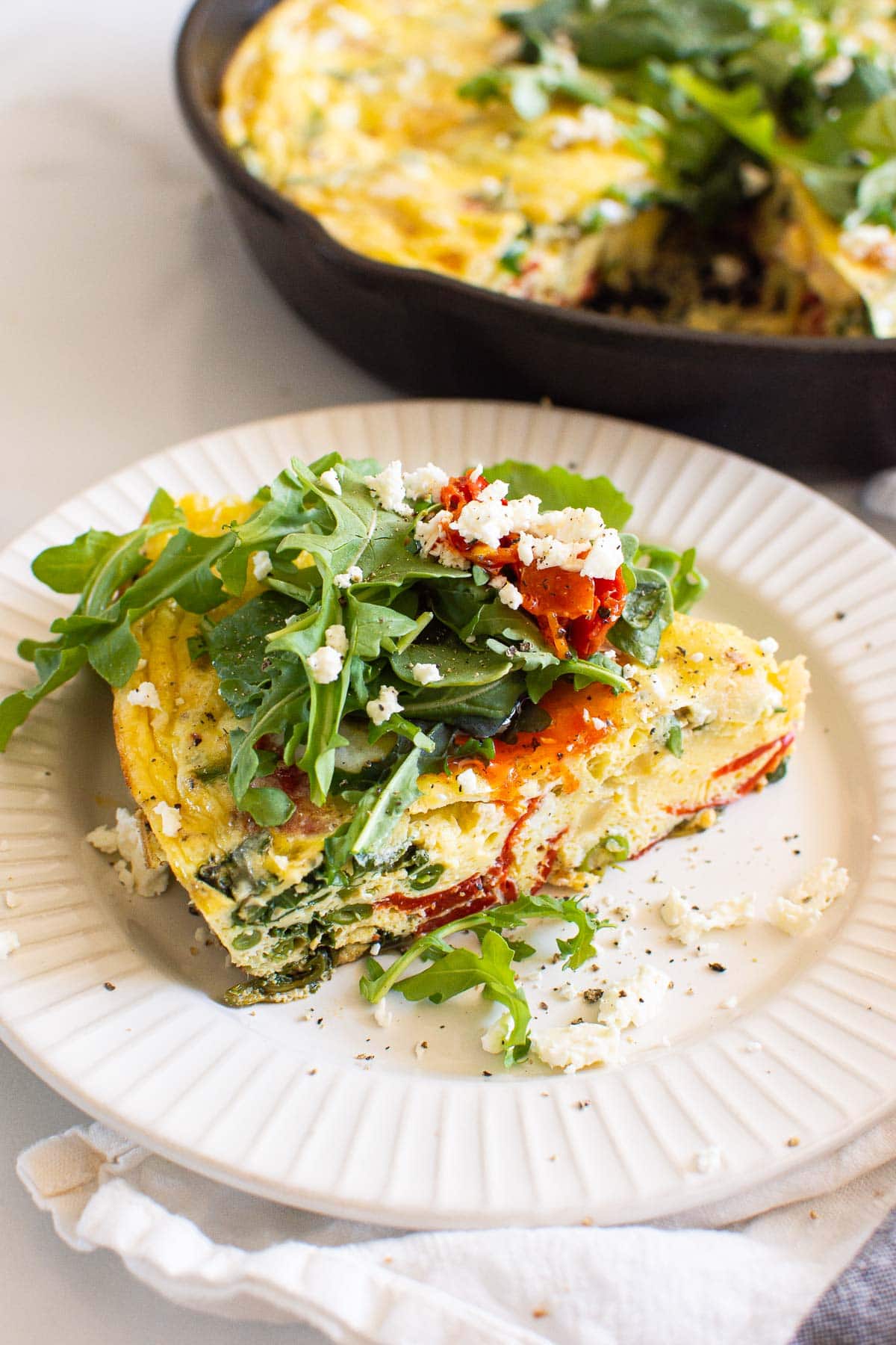 A slice of frittata with red pepper, greens and feta cheese garnish on a plate.