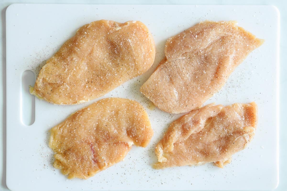 Four seasoned chicken breast cutlets on cutting board.