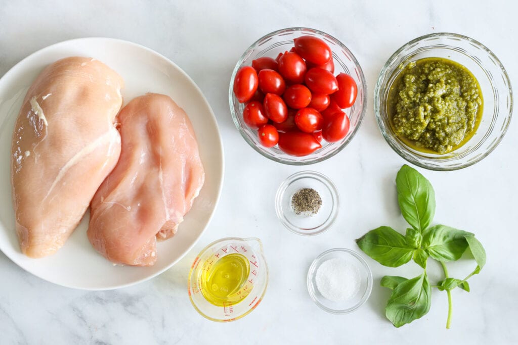 Ingredients for Pesto Chicken, including check breasts, cherry tomatoes, basil pesto, basil, salt, pepper, and olive oil.