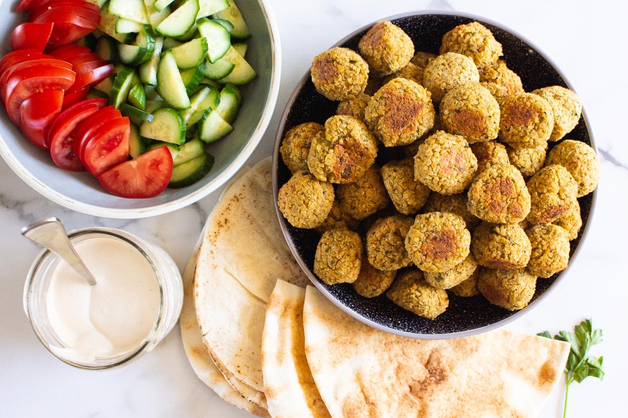 falafel in a bowl pitas on plate with tahini sauce and chopped cucumber tomatoes in a bowl