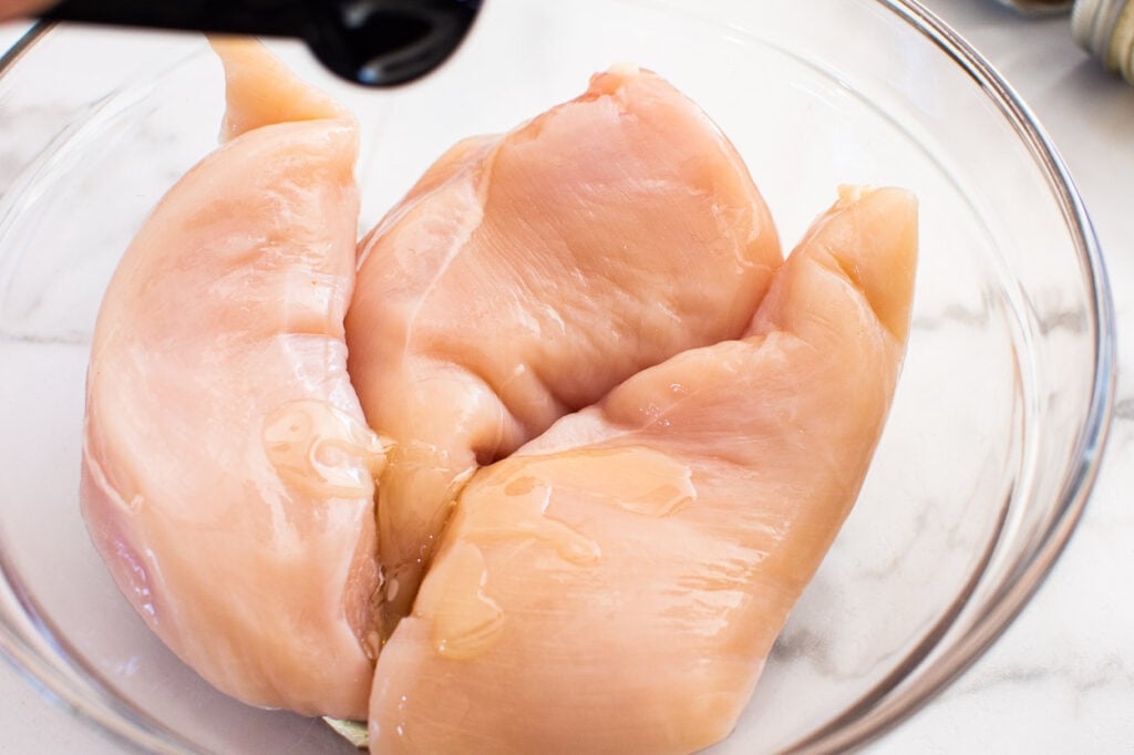 Person drizzling oil on top of three chicken breasts in a bowl.