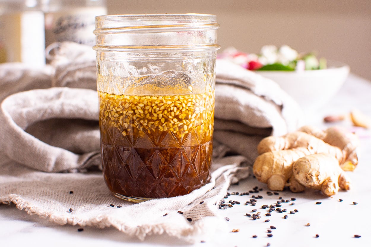 Homemade Asian salad dressing in mason jar with ginger knob and napkin and sesame seeds on a counter.