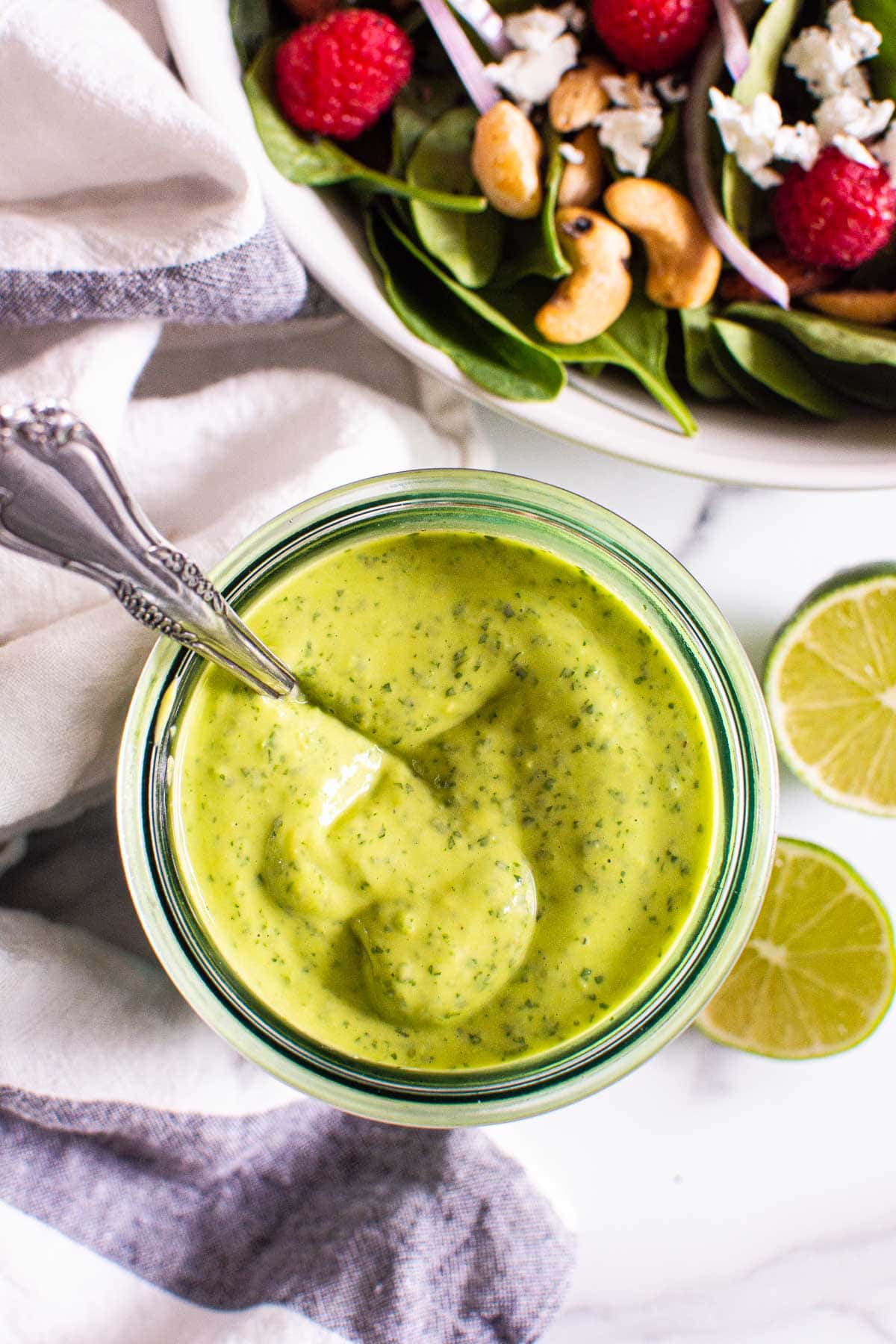 Avocado cilantro dressing in a glass jar with a spoon and a bowl of salad and limes nearby.