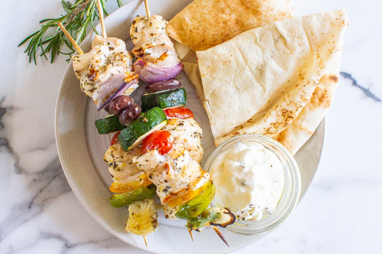 Greek chicken kabobs on plate with sauce in a bowl and pita.