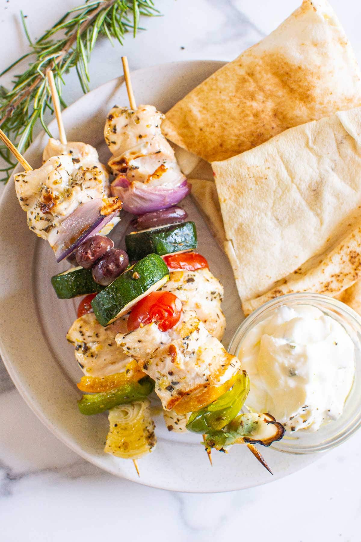 Three Greek chicken kabobs on plate with pita and dip.