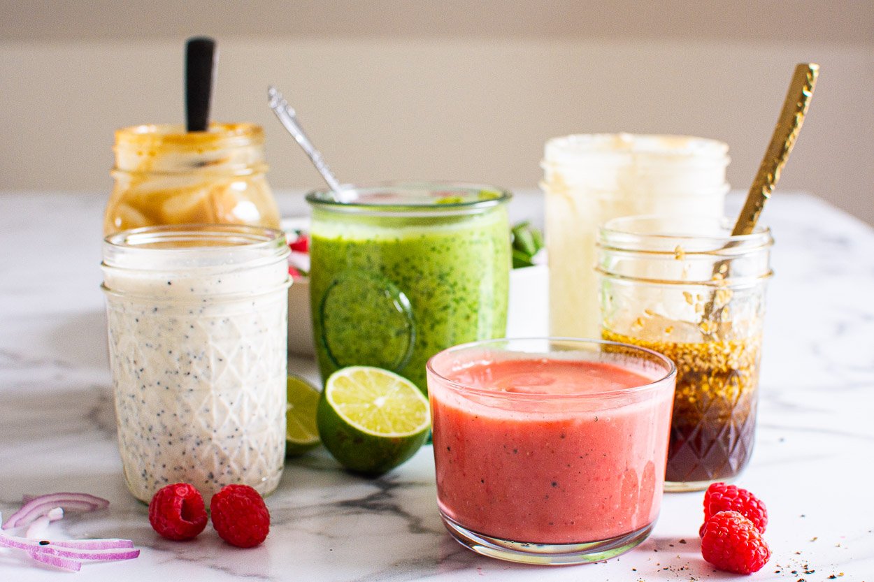 Six healthy homemade salad dressings in mason jars with spoons.