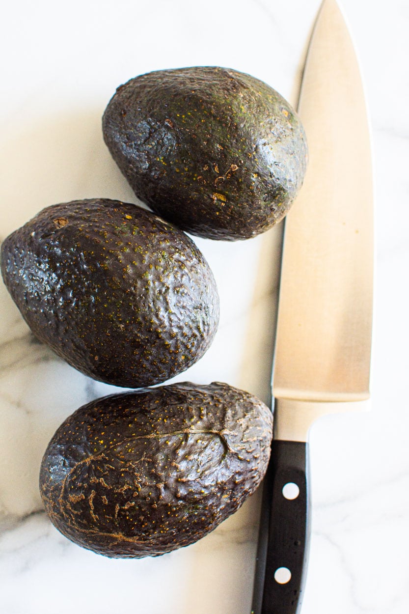Three avocados and a chef's knife on a countertop.