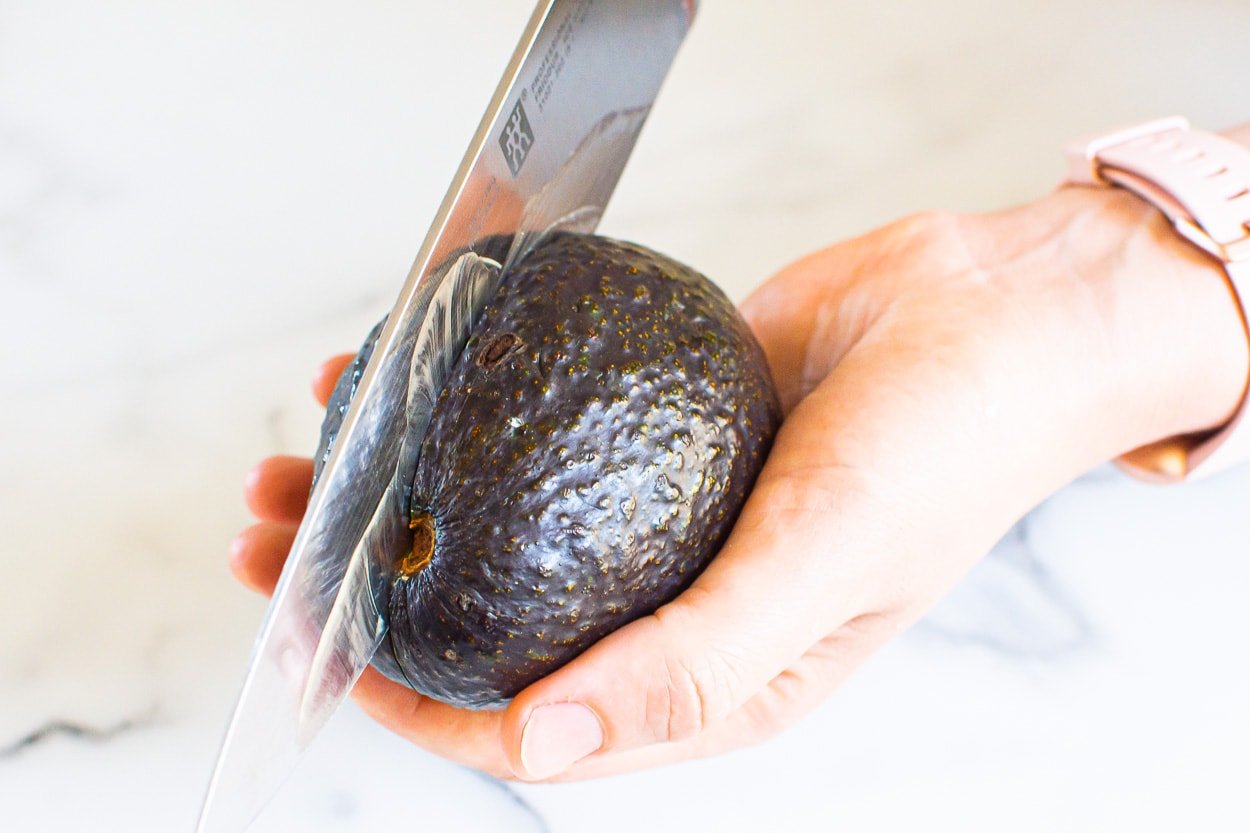 Person holding avocado in hand and slicing it in half with a knife.