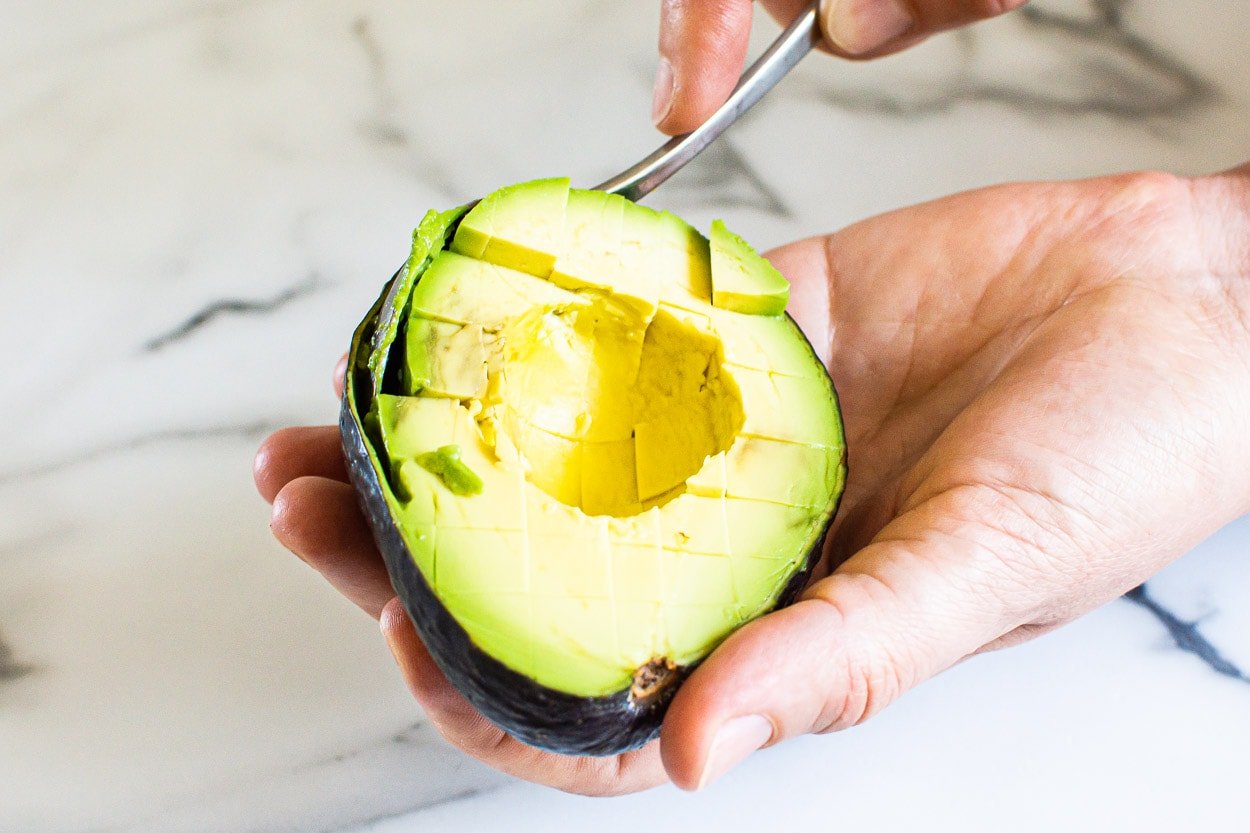 Person scooping out diced avocado with a tablespoon from its shell.