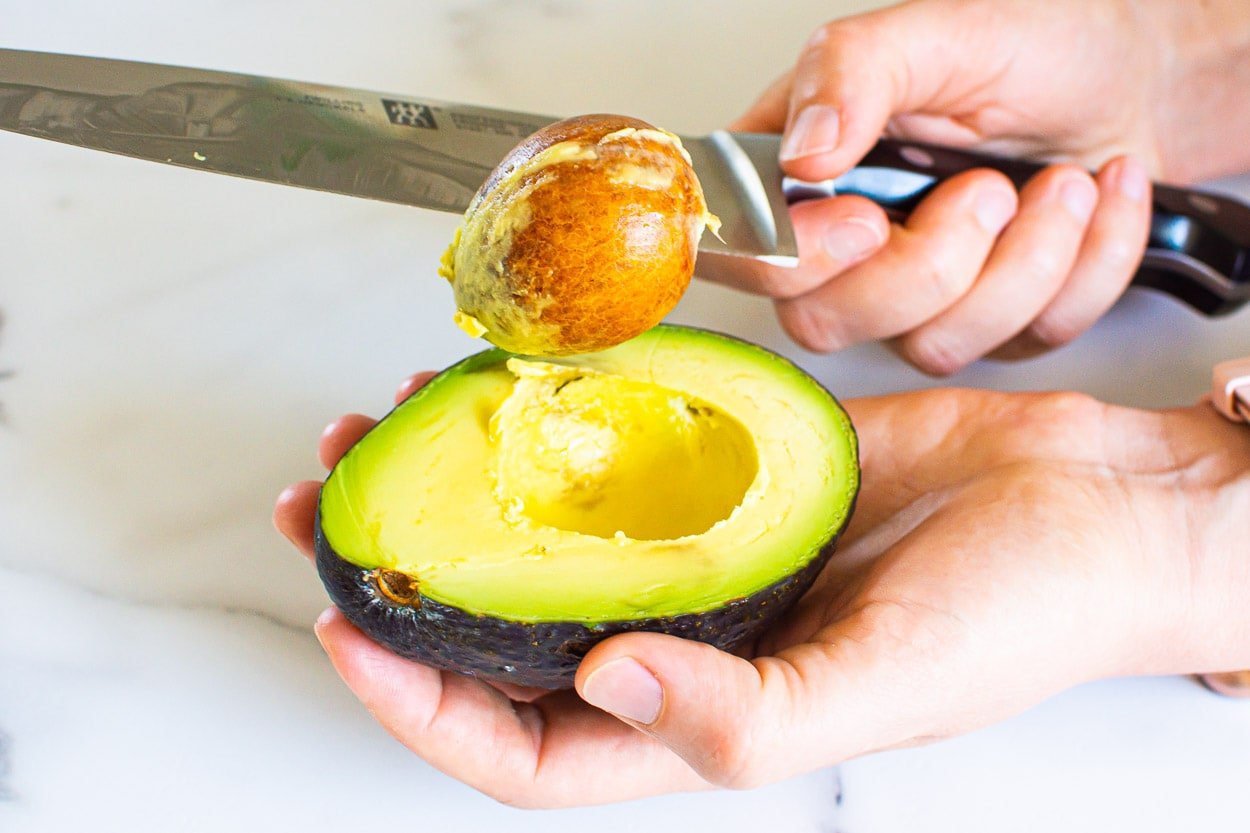 Person removing a pit from an avocado half with a knife.