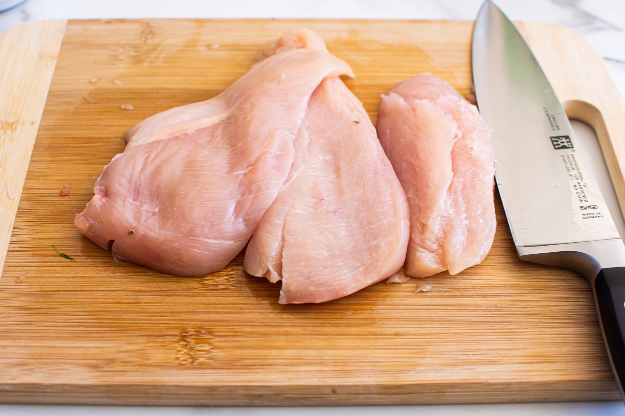 Sliced chicken breasts in half on a cutting board with a knife.