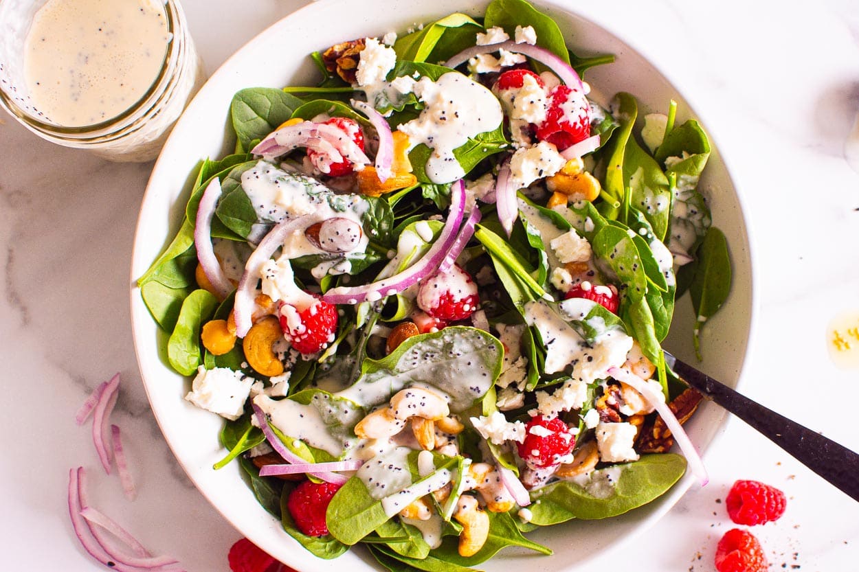 Poppyseed dressing drizzled on salad with raspberries and nuts in a bowl. Jar with the dressing near it.