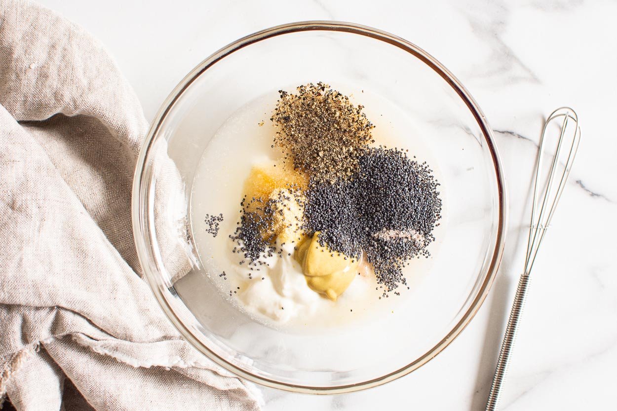 Greek yogurt, maple syrup, poppy seeds, apple cider vinegar, dijon mustard, onion powder, salt and pepper in a bowl with a whisk.