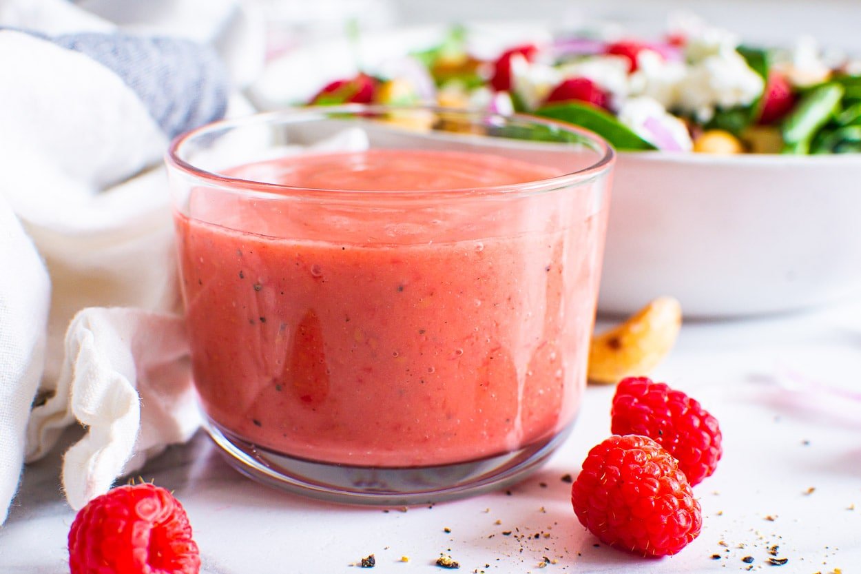 Raspberry vinaigrette in a small glass jar with a salad behind it.