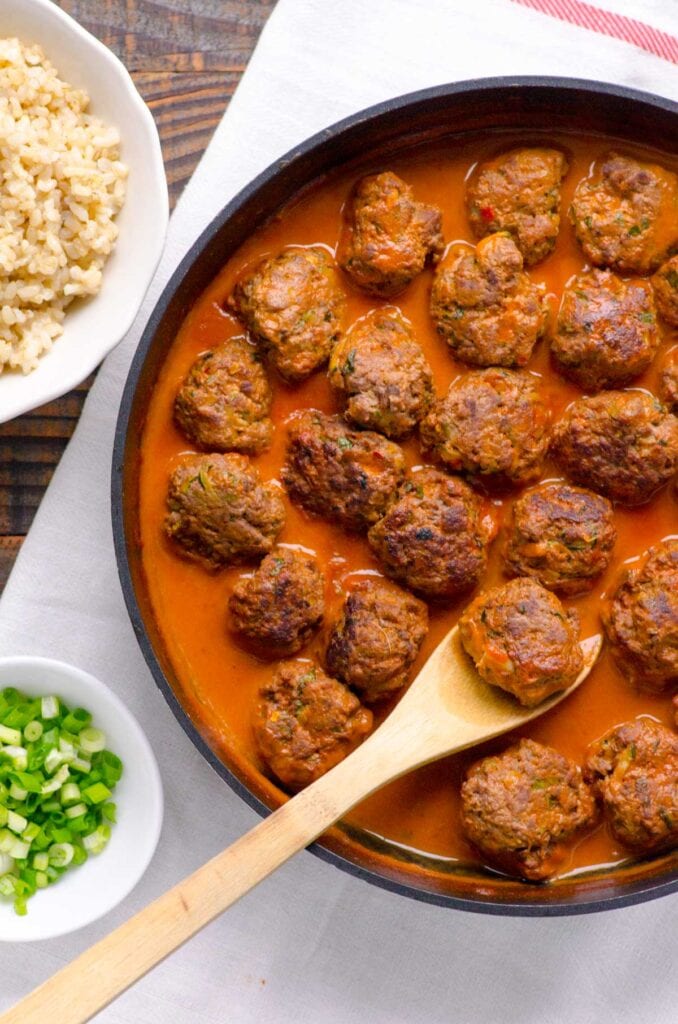 Thai meatballs in large skillet with serving spoon, a bowl of rice and bowl of garnish.