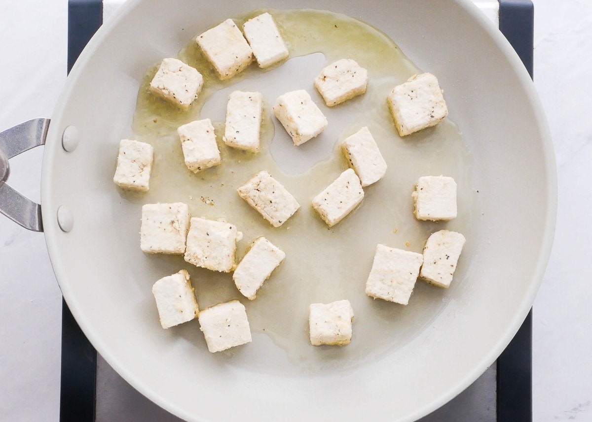 Cubed tofu being fried in white skillet in oil.