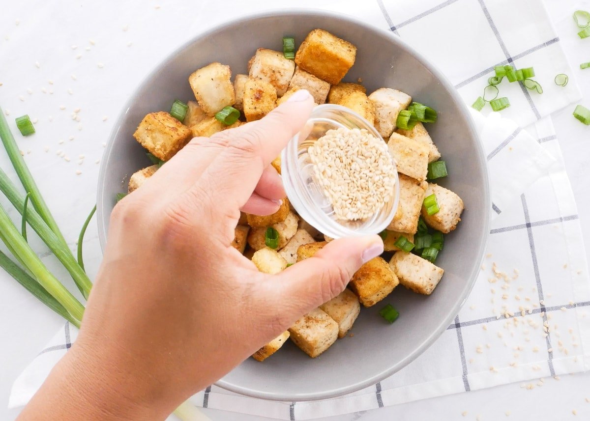 Sprinkling fried tofu in a bowl with sesame seeds and chopped green onion.