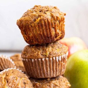 Three healthy apple muffins stacked on top of each other. Apples, cinnamon sticks and napkin on a counter.