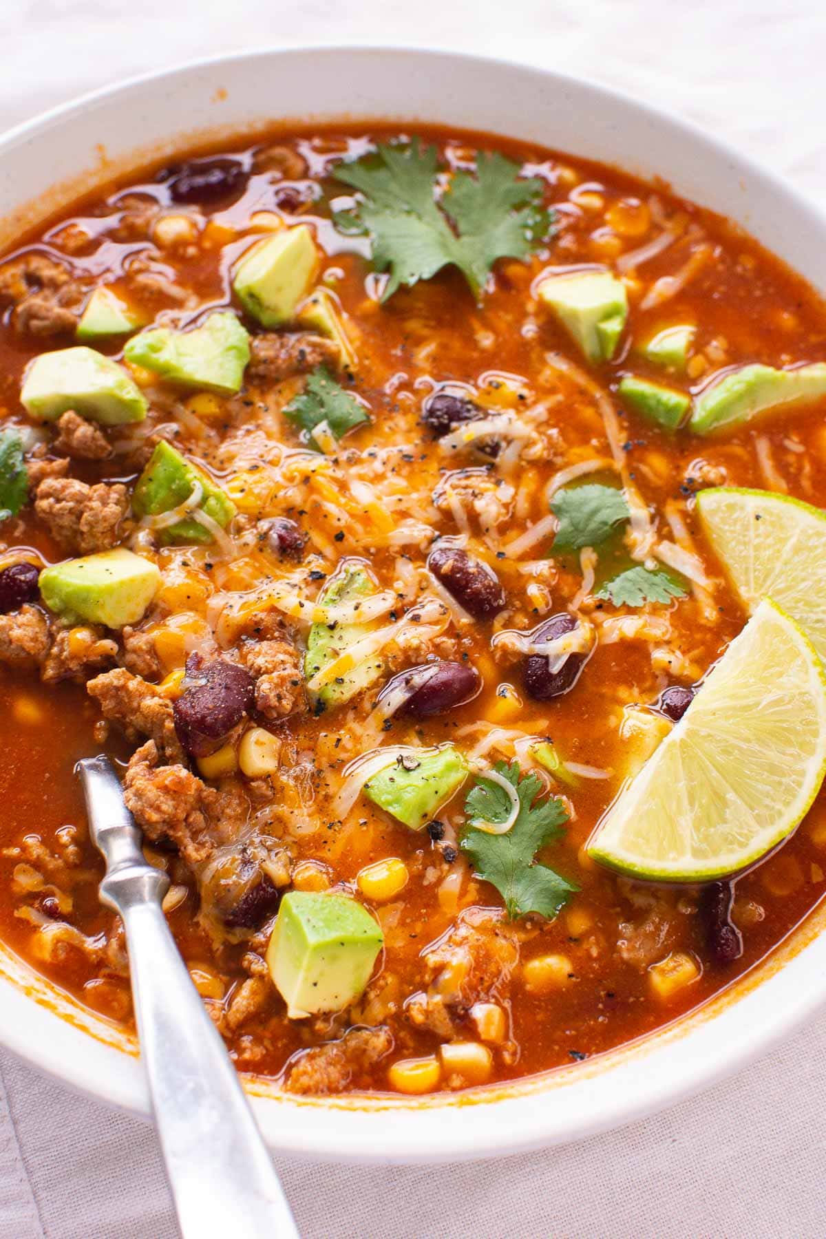 Ground turkey taco soup in white bowl with spoon and lime, avocado and cilantro garnish.