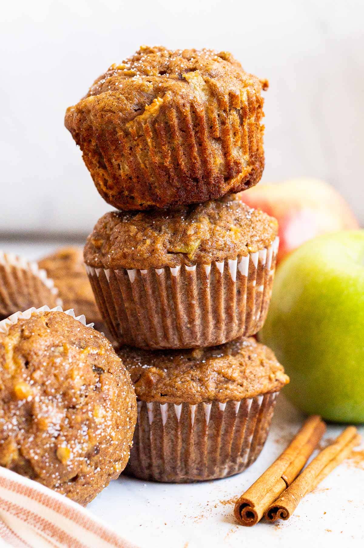 Three healthy apple muffins stacked on top of each other. Apples, cinnamon sticks and napkin on a counter.