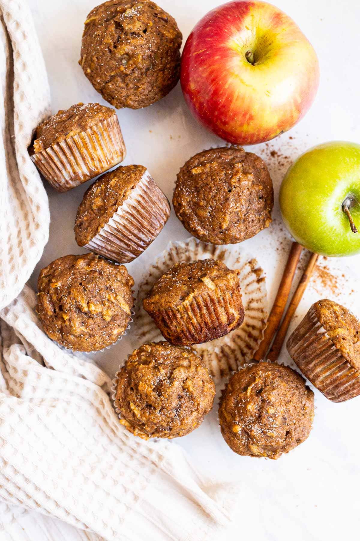 Nine healthy apple muffins sprinkled with sugar. One muffin unwrapped in the center. Decor around.