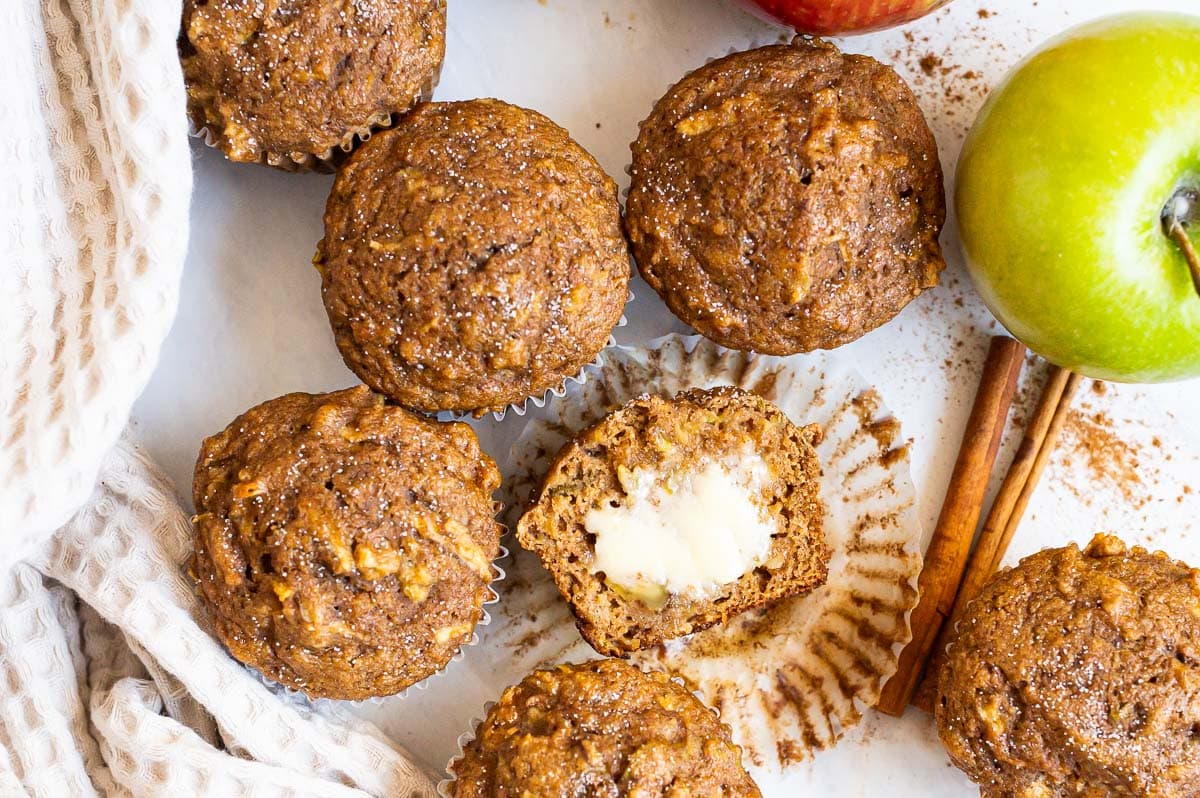 One apple muffin sliced in half with butter. More muffins, green apple, napkin on a counter around it.