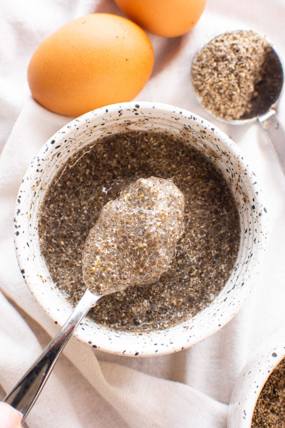 Chia egg on a spoon over white bowl, eggs and ground chia in measuring cup nearby.