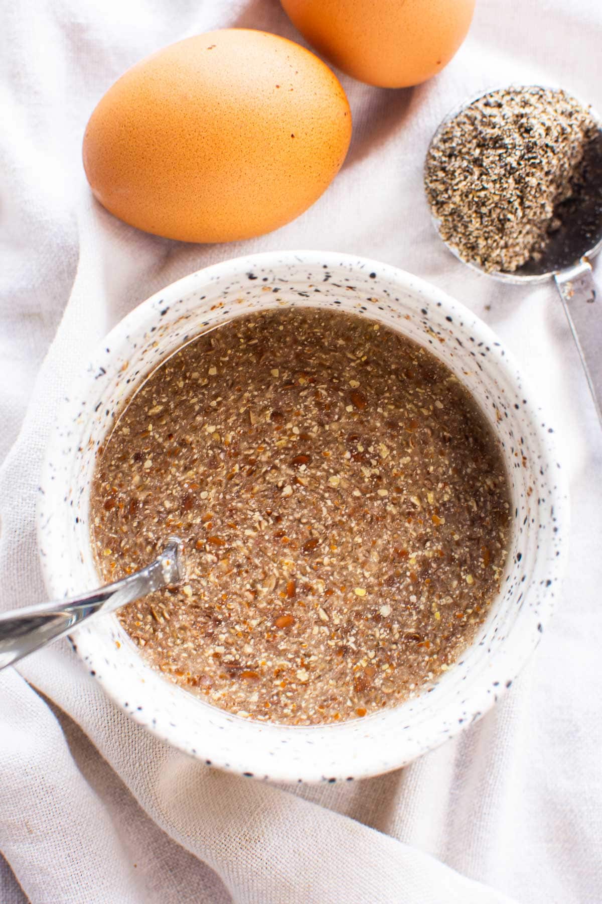 Flax egg in a bowl with a spoon. Two brown eggs and ground flaxseed in measuring spoon.