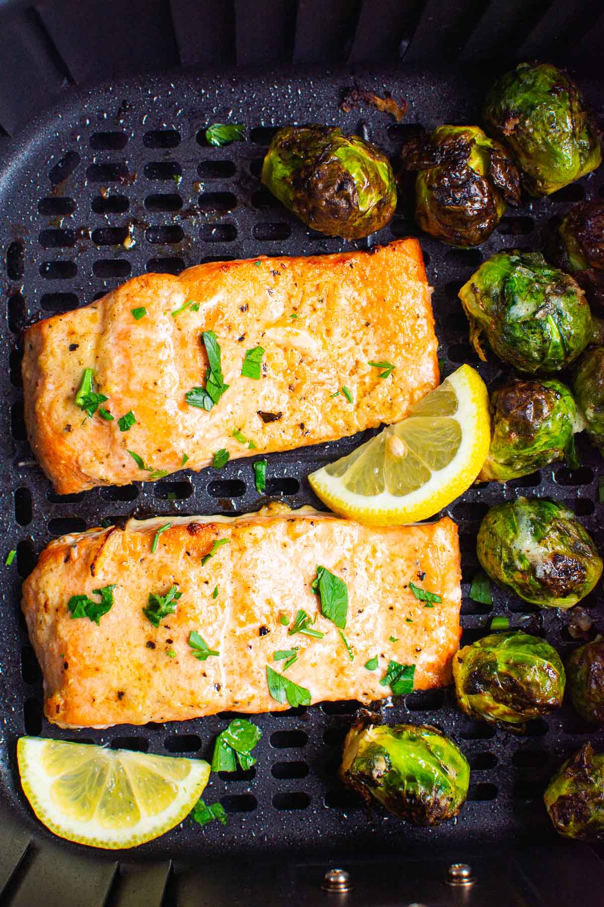 Air fryer salmon with brussels sprouts in air fryer basket with parsley and lemon wedges.