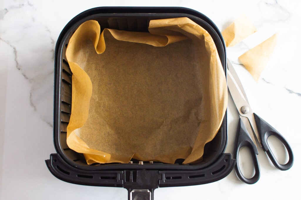 Air fryer basket lined with parchment paper and scissors laying on the counter.