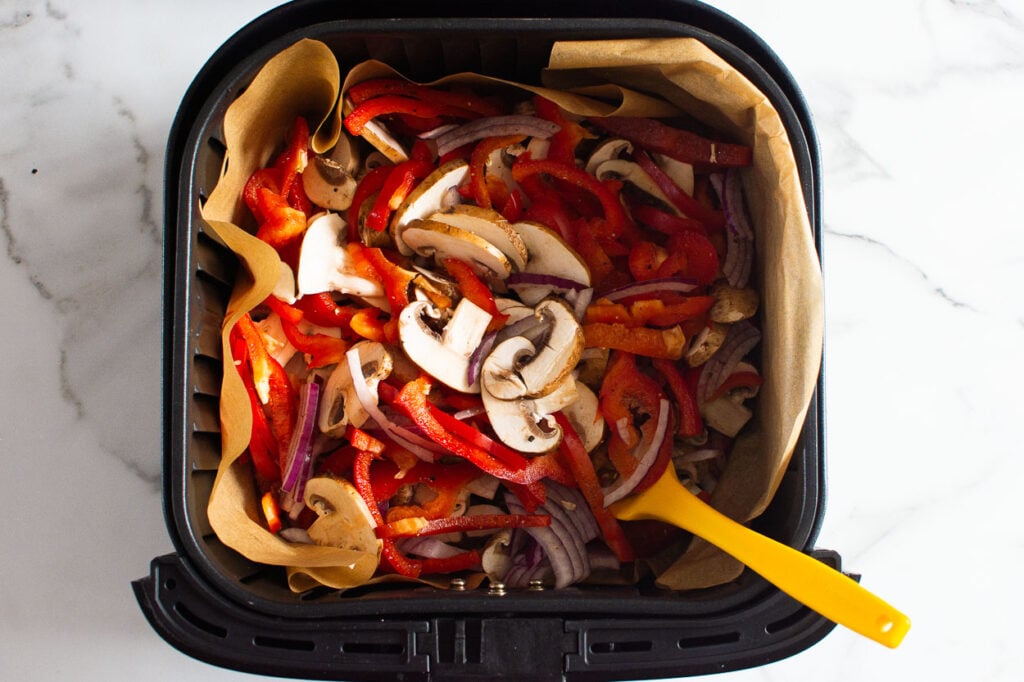 Mushrooms, red pepper and red onion in air fryer basket mixed with yellow spatula.