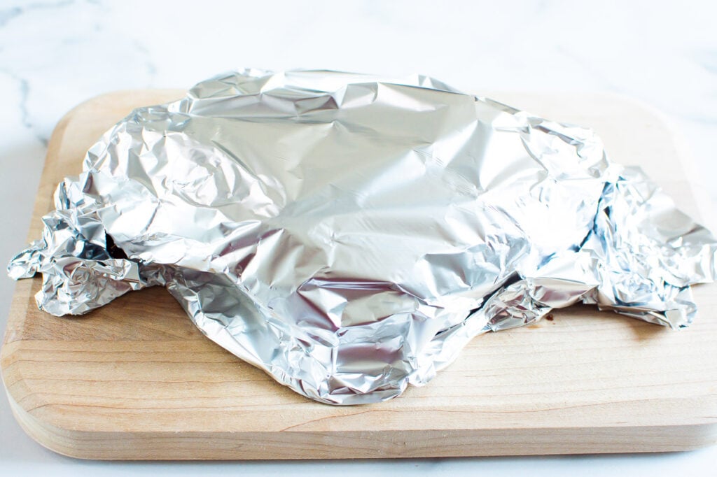 Cooked turkey breast with aluminum foil over it on cutting board.