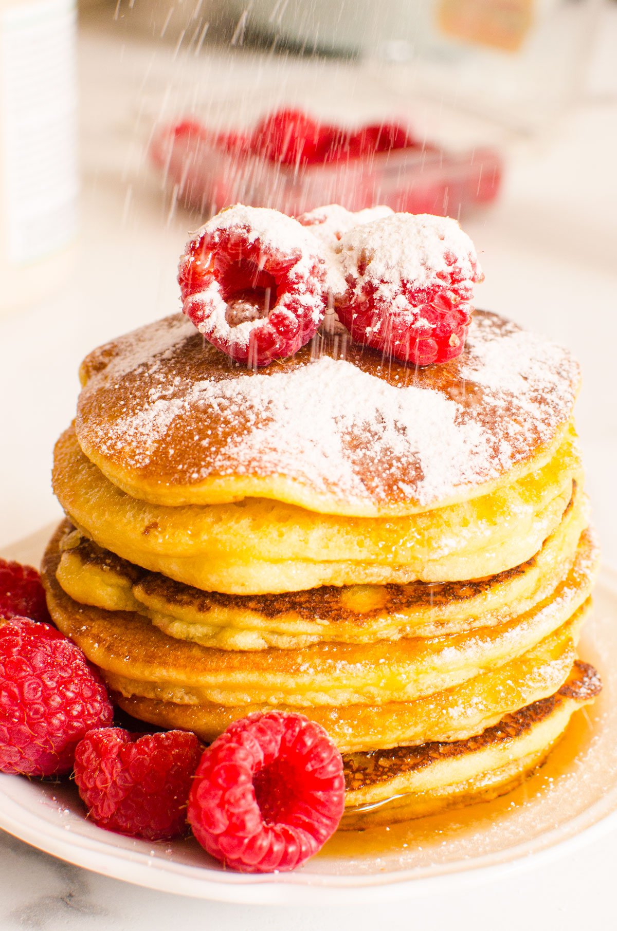 Stack of almond flour pancakes with raspberries, maple syrup and icing sugar on a plate.