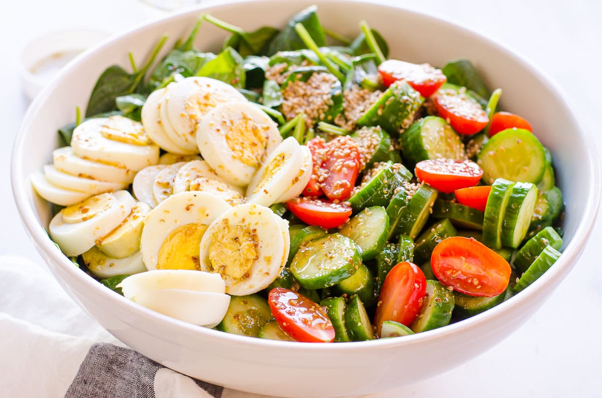 Spinach salad with hard boiled eggs, cucumbers and tomatoes dressed in a white bowl.