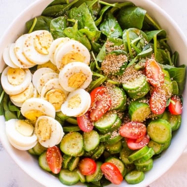 A bowl of best spinach salad with hard boiled eggs, tomatoes, cucumbers and sesame seeds.