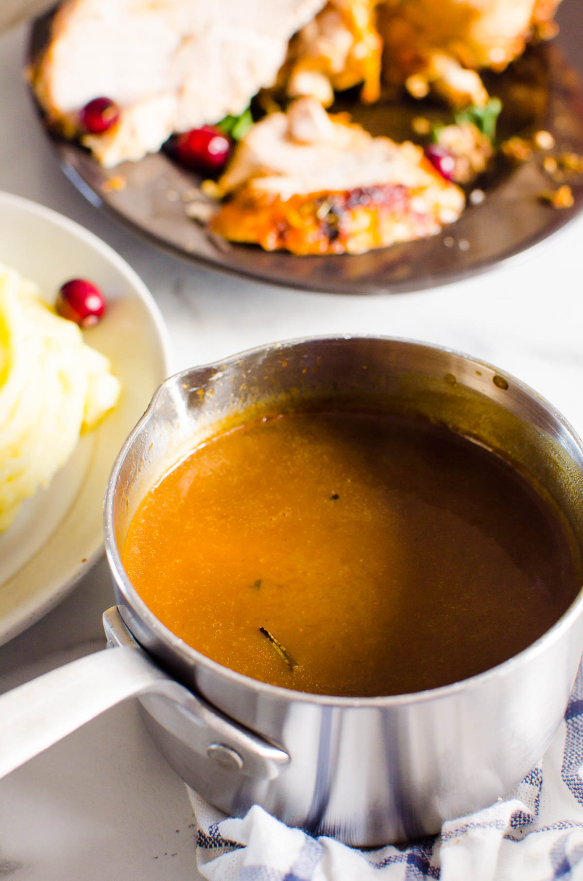 Turkey gravy in small pot, mashed potatoes and turkey in serving dishes nearby.